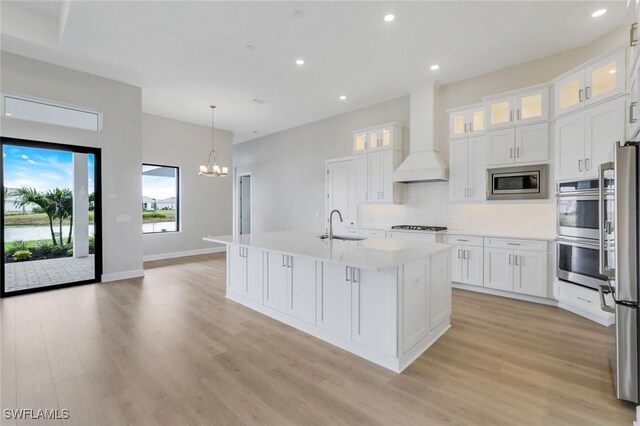 kitchen featuring light wood finished floors, a sink, premium range hood, a chandelier, and stainless steel appliances