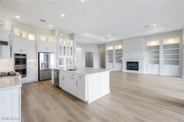 kitchen with appliances with stainless steel finishes, an island with sink, a sink, white cabinets, and light wood-type flooring