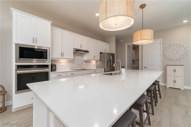 kitchen with under cabinet range hood, a sink, backsplash, stainless steel appliances, and a large island with sink