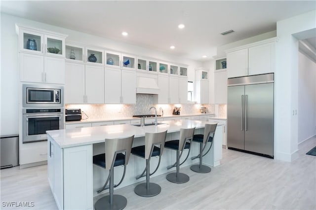 kitchen with custom range hood, light countertops, built in appliances, and an island with sink