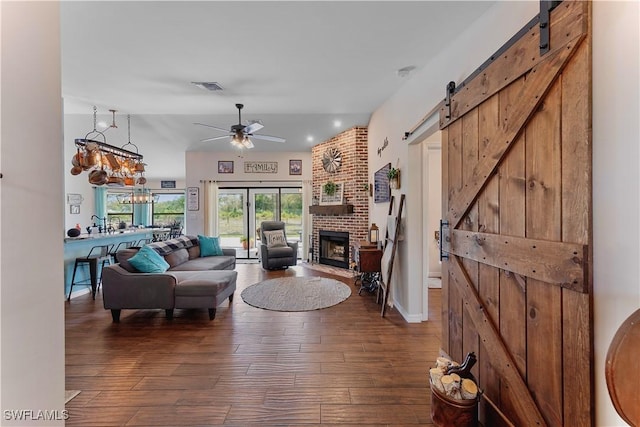 living room with ceiling fan with notable chandelier, a barn door, visible vents, a brick fireplace, and dark wood-style floors