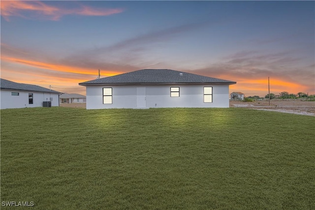 back of house at dusk featuring a shingled roof, a yard, stucco siding, and central AC unit