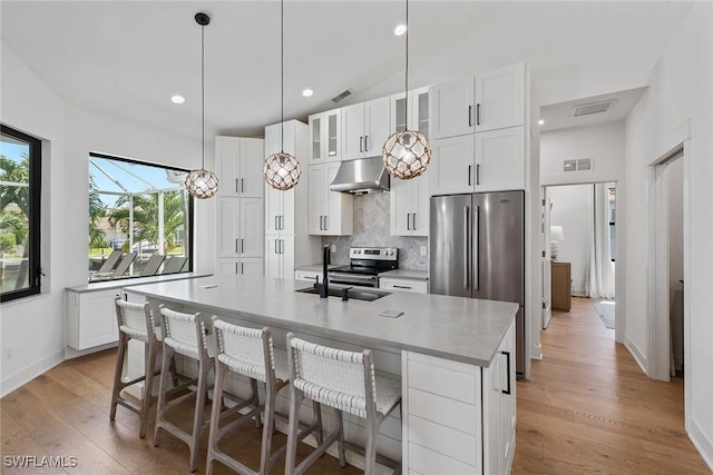 kitchen with appliances with stainless steel finishes, under cabinet range hood, backsplash, light wood finished floors, and a center island with sink