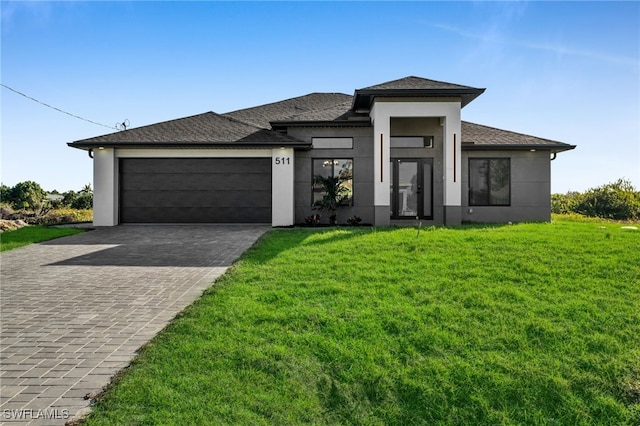 prairie-style home with a garage, stucco siding, a front lawn, a shingled roof, and decorative driveway