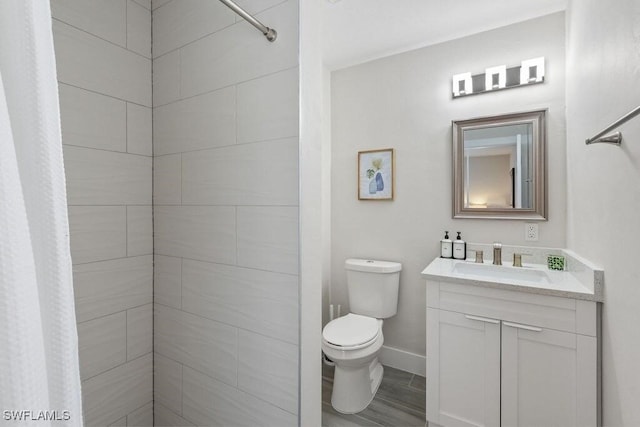bathroom with toilet, tiled shower, vanity, and baseboards