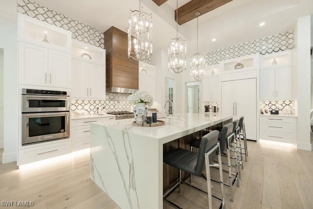 6145 Sea Grass Ln, Naples, FL 34116 - kitchen with stainless steel double oven, premium range hood, tasteful backsplash, beamed ceiling, and light wood finished floors