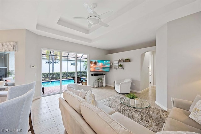 living room with light tile patterned floors, a ceiling fan, arched walkways, a raised ceiling, and baseboards