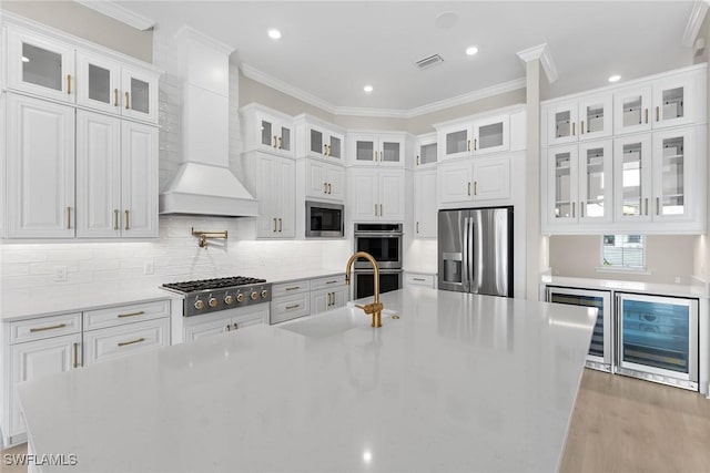 kitchen with stainless steel appliances, wine cooler, wall chimney range hood, crown molding, and an island with sink