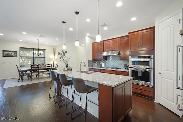 kitchen with a sink, under cabinet range hood, dark wood-style floors, light stone countertops, and backsplash