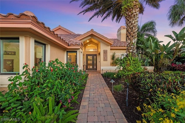 exterior entry at dusk featuring stucco siding, a chimney, a tile roof, and french doors