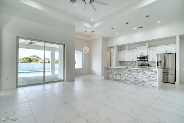 unfurnished living room with a high ceiling, ceiling fan, a tray ceiling, light marble finish floors, and a sink