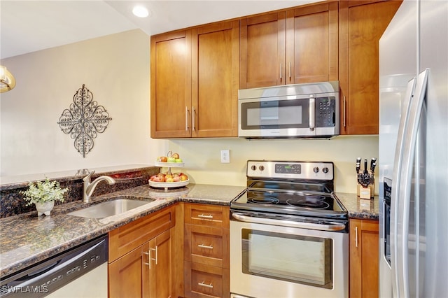 kitchen featuring appliances with stainless steel finishes, a sink, dark stone counters, brown cabinetry, and recessed lighting