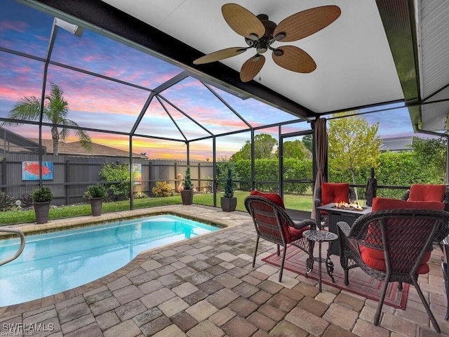 pool at dusk with an outdoor fire pit, a patio, glass enclosure, fence, and ceiling fan