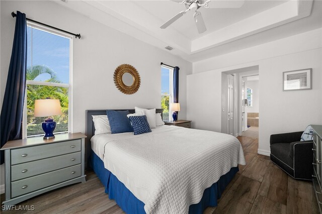 bedroom featuring a tray ceiling, wood finished floors, multiple windows, and ensuite bath