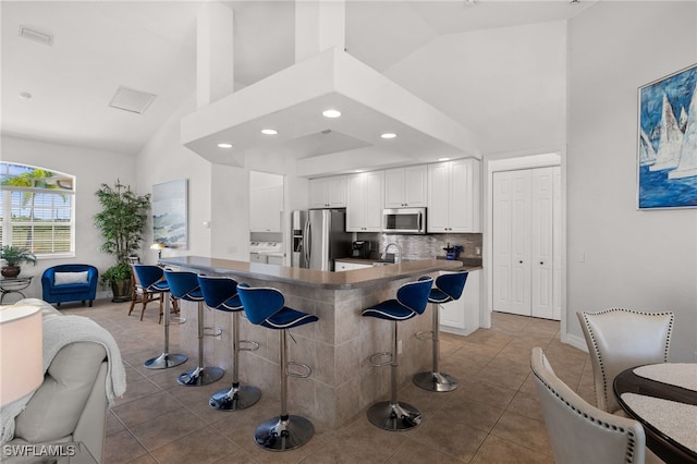 kitchen featuring stainless steel appliances, high vaulted ceiling, a kitchen bar, tile patterned flooring, and white cabinetry