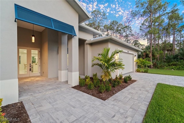 view of side of property with french doors, stucco siding, an attached garage, decorative driveway, and a lawn