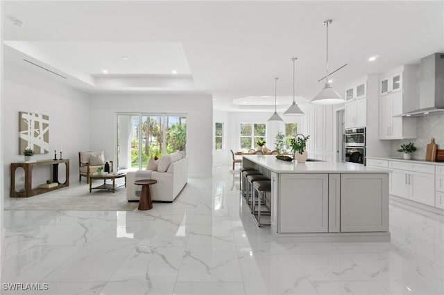 kitchen with wall chimney range hood, stainless steel double oven, a tray ceiling, light countertops, and white cabinetry