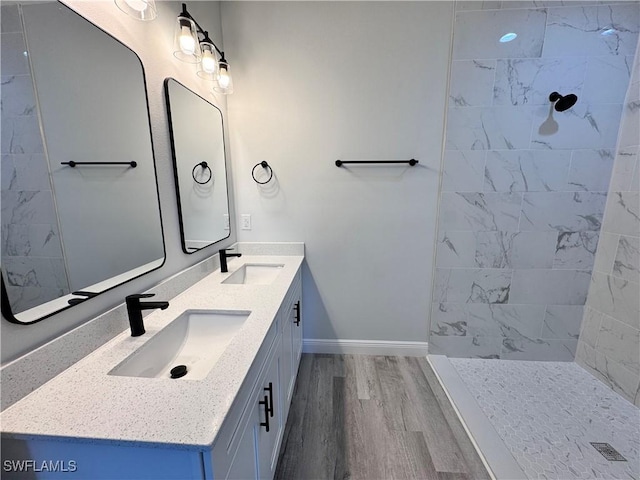 full bathroom with wood finished floors, baseboards, double vanity, and tiled shower
