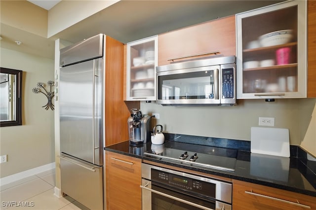 kitchen featuring stainless steel appliances, light tile patterned floors, dark stone countertops, glass insert cabinets, and brown cabinets