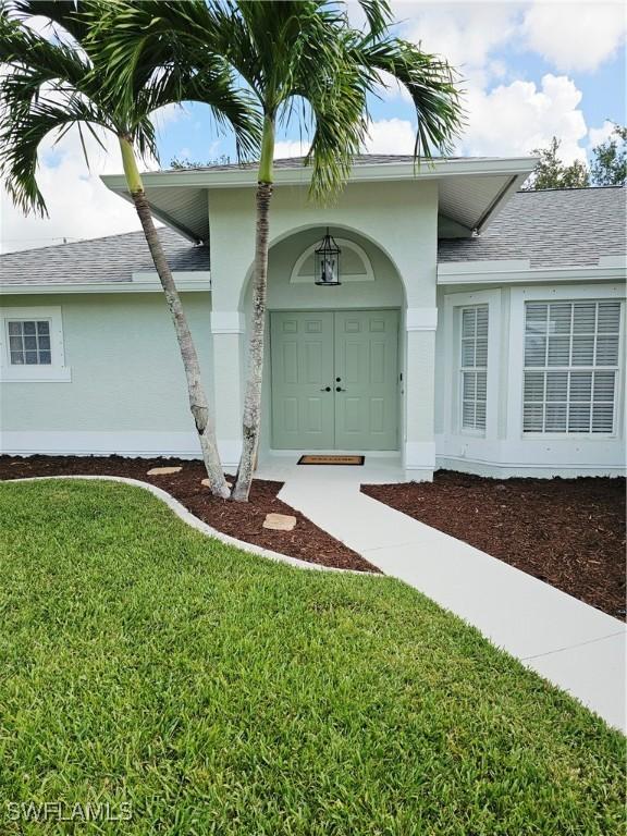1221 SW 10th Ter, Cape Coral, FL 33991 - property entrance with stucco siding, a yard, and a shingled roof