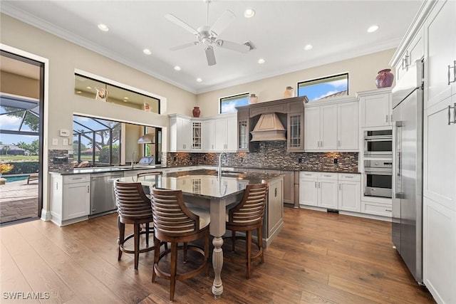 11950 Heather Woods Ct, Naples, FL 34120 - kitchen with stainless steel appliances, custom exhaust hood, ornamental molding, a kitchen island with sink, and a ceiling fan