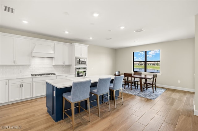 20676 Napa Loop, Estero, FL 33928 - kitchen with stainless steel appliances, premium range hood, a kitchen island with sink, light wood-style floors, and white cabinetry