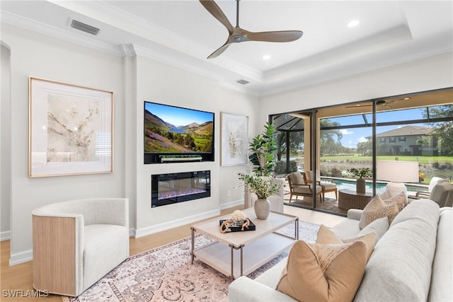 8834 Vaccaro Ct, Naples, FL 34119 - living room featuring a ceiling fan, crown molding, a raised ceiling, a glass covered fireplace, and light wood-style floors