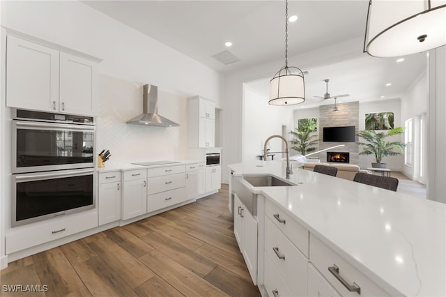 2274 Hawksridge Dr, Naples, FL 34105 - kitchen with wall chimney exhaust hood, double oven, ceiling fan, a fireplace, and black electric cooktop