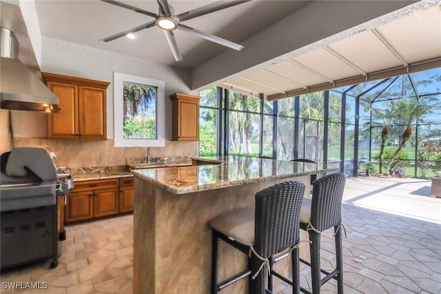4323 Silver Fox Dr, Naples, FL 34119 - kitchen with wall chimney exhaust hood, brown cabinetry, a ceiling fan, tasteful backsplash, and a kitchen bar