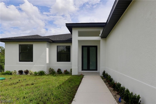 2713 8th St W, Lehigh Acres, FL 33971 - view of exterior entry featuring stucco siding, a yard, and a shingled roof