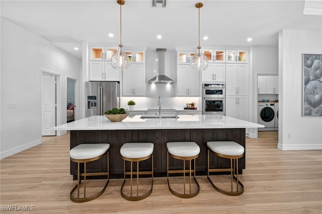 7637 Mulberry Ln, Naples, FL 34114 - kitchen with wall chimney exhaust hood, appliances with stainless steel finishes, light wood-style flooring, a large island with sink, and light countertops