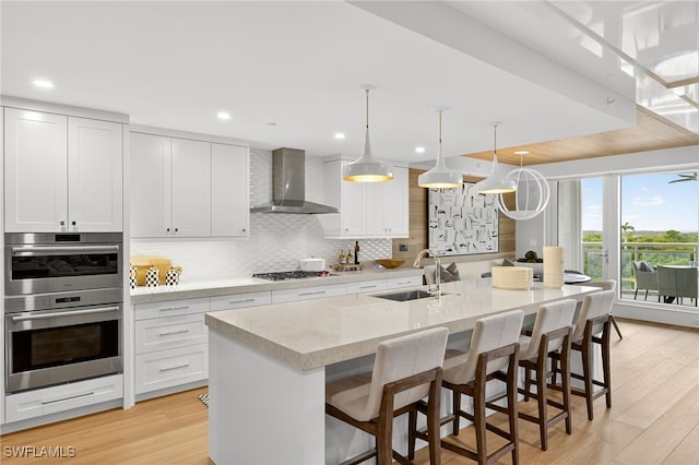 13925 Old Coast Rd Unit 403, Naples, FL 34110 - kitchen featuring wall chimney range hood, double oven, tasteful backsplash, white cabinets, and light wood-style flooring