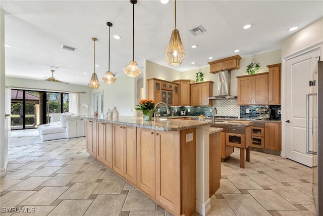 3917 La Vida Way, Cape Coral, FL 33993 - kitchen featuring wall chimney range hood, an island with sink, brown cabinetry, decorative backsplash, and light stone counters