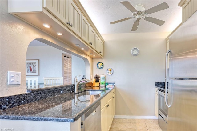 kitchen featuring stainless steel appliances, light tile flooring, ceiling fan, dark stone countertops, and sink