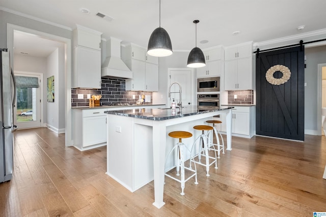 kitchen featuring stainless steel appliances, a barn door, light hardwood / wood-style floors, an island with sink, and premium range hood