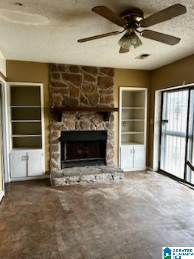 unfurnished living room featuring ceiling fan, a stone fireplace, built in shelves, and a textured ceiling