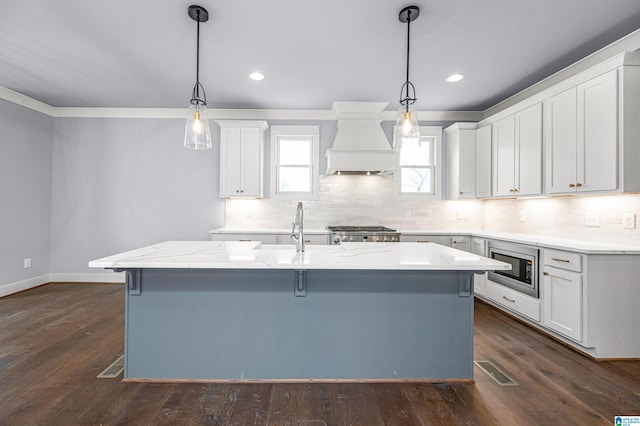 kitchen featuring dark wood-type flooring, stainless steel microwave, an island with sink, and custom range hood