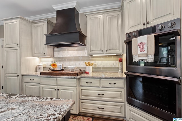 kitchen featuring light stone counters, double oven, decorative backsplash, custom exhaust hood, and ornamental molding