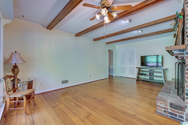 living room featuring beamed ceiling, light hardwood / wood-style floors, a brick fireplace, and ceiling fan