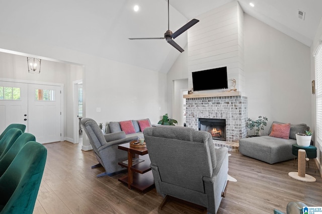 living room featuring high vaulted ceiling, wood-type flooring, a brick fireplace, and ceiling fan with notable chandelier