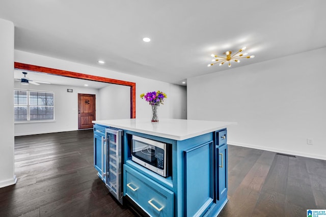 kitchen with ceiling fan with notable chandelier, beverage cooler, blue cabinetry, dark wood-type flooring, and stainless steel microwave