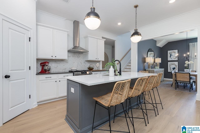 kitchen with wall chimney range hood, backsplash, hanging light fixtures, light hardwood / wood-style flooring, and a kitchen island with sink