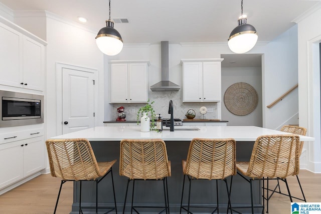 kitchen featuring wall chimney exhaust hood, a kitchen island with sink, light wood-type flooring, backsplash, and stainless steel microwave