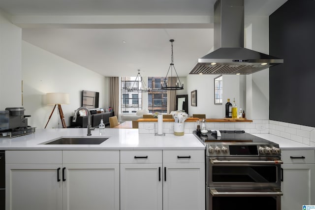 kitchen with wall chimney range hood, white cabinetry, hanging light fixtures, range with two ovens, and sink