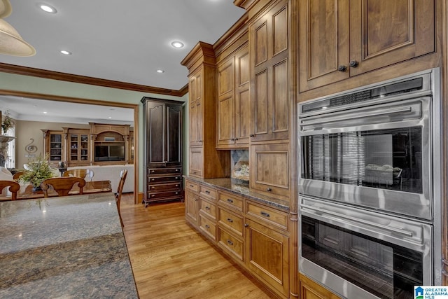 kitchen featuring ornamental molding, dark stone countertops, stainless steel double oven, and light hardwood / wood-style flooring