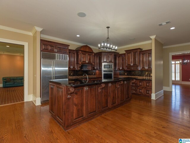 kitchen with dark hardwood / wood-style floors, tasteful backsplash, built in appliances, an inviting chandelier, and a kitchen island with sink