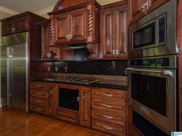 kitchen with dark stone counters, light hardwood / wood-style floors, backsplash, built in appliances, and ornamental molding