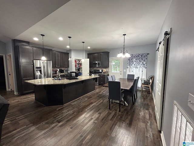 kitchen with a barn door, a center island with sink, decorative light fixtures, stainless steel appliances, and dark hardwood / wood-style flooring