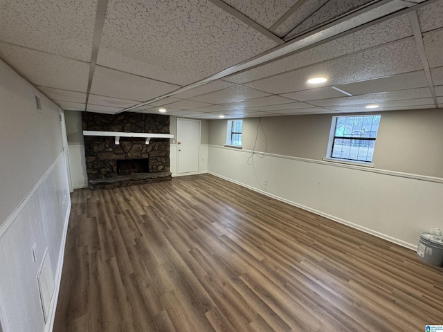 basement featuring a wainscoted wall, a drop ceiling, a stone fireplace, and wood finished floors