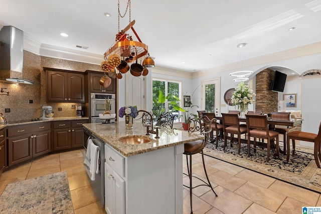 kitchen with sink, decorative backsplash, dark brown cabinets, a center island with sink, and wall chimney exhaust hood